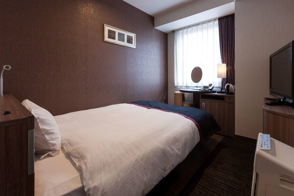 Et hotelværelse med en seng, et skrivebord med lampe, et fjernsyn og et vindue med gardiner.