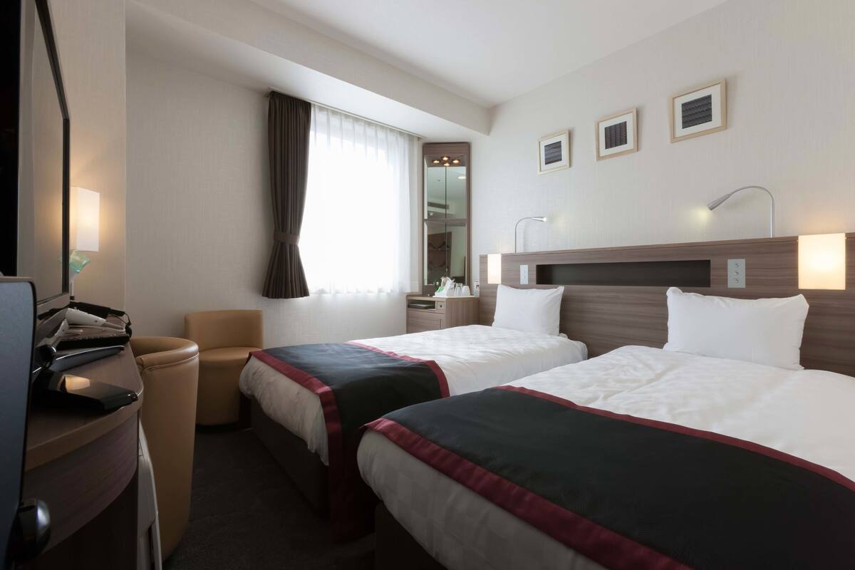 Et hotelværelse med to senge, et skrivebord, en stol, et fjernsyn og et vindue med gardiner.