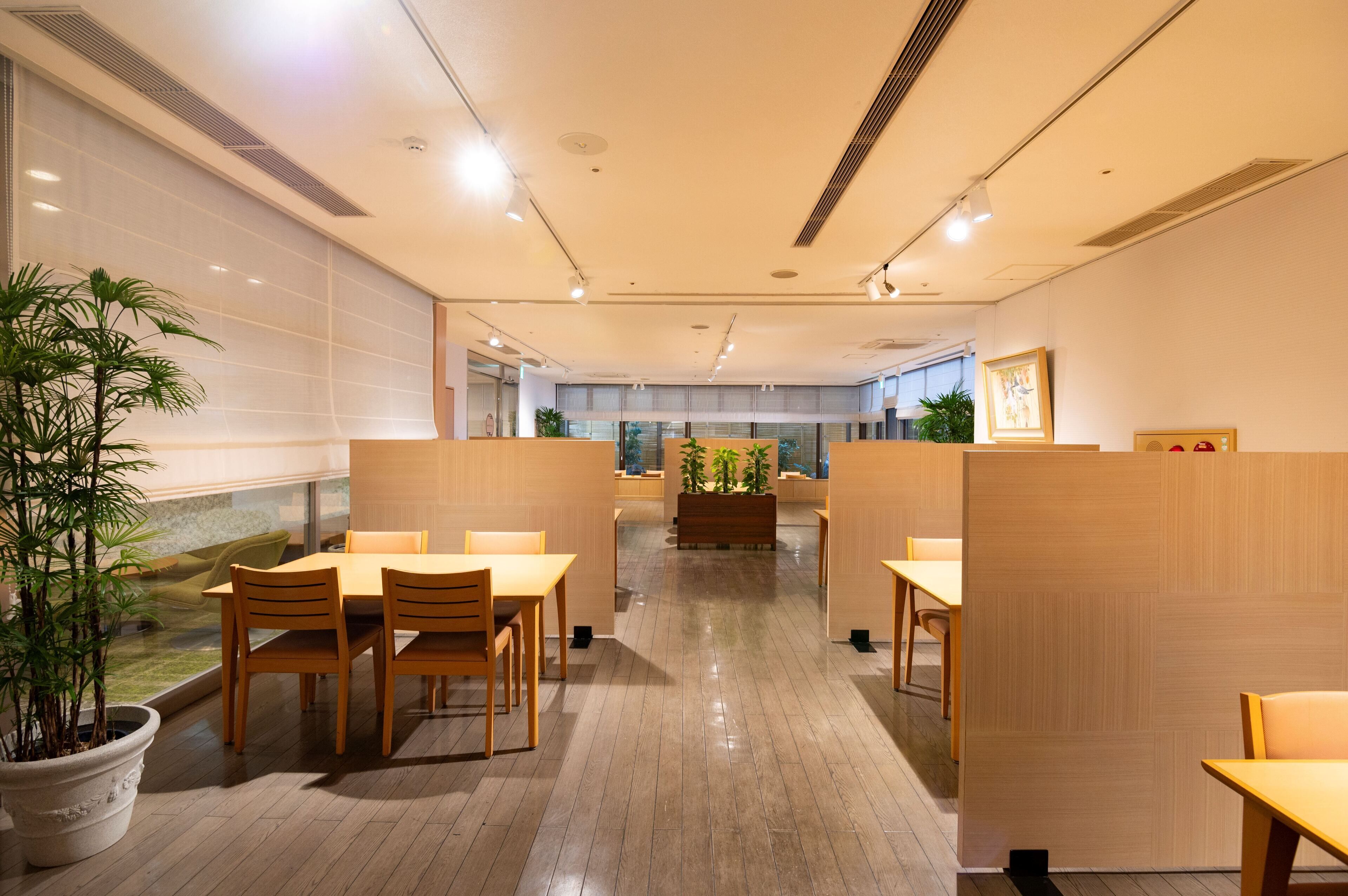 A modern office space with wooden tables and chairs, potted plants, and a view of the city through large windows.