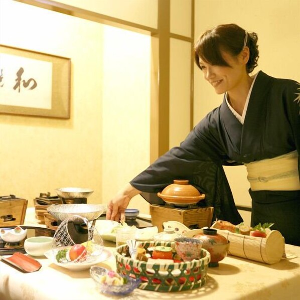 Une personne vêtue d’un costume traditionnel servant des plats à une table où se trouvent divers mets et ustensiles.