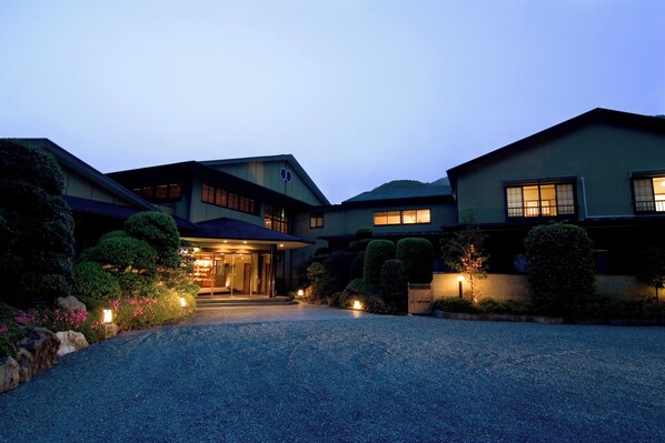 A multi-story building with a well-lit entrance, surrounded by landscaped gardens and a gravel driveway. - Hotel Kawachiya Izu Shimogamo Onsen (Minamiizu)