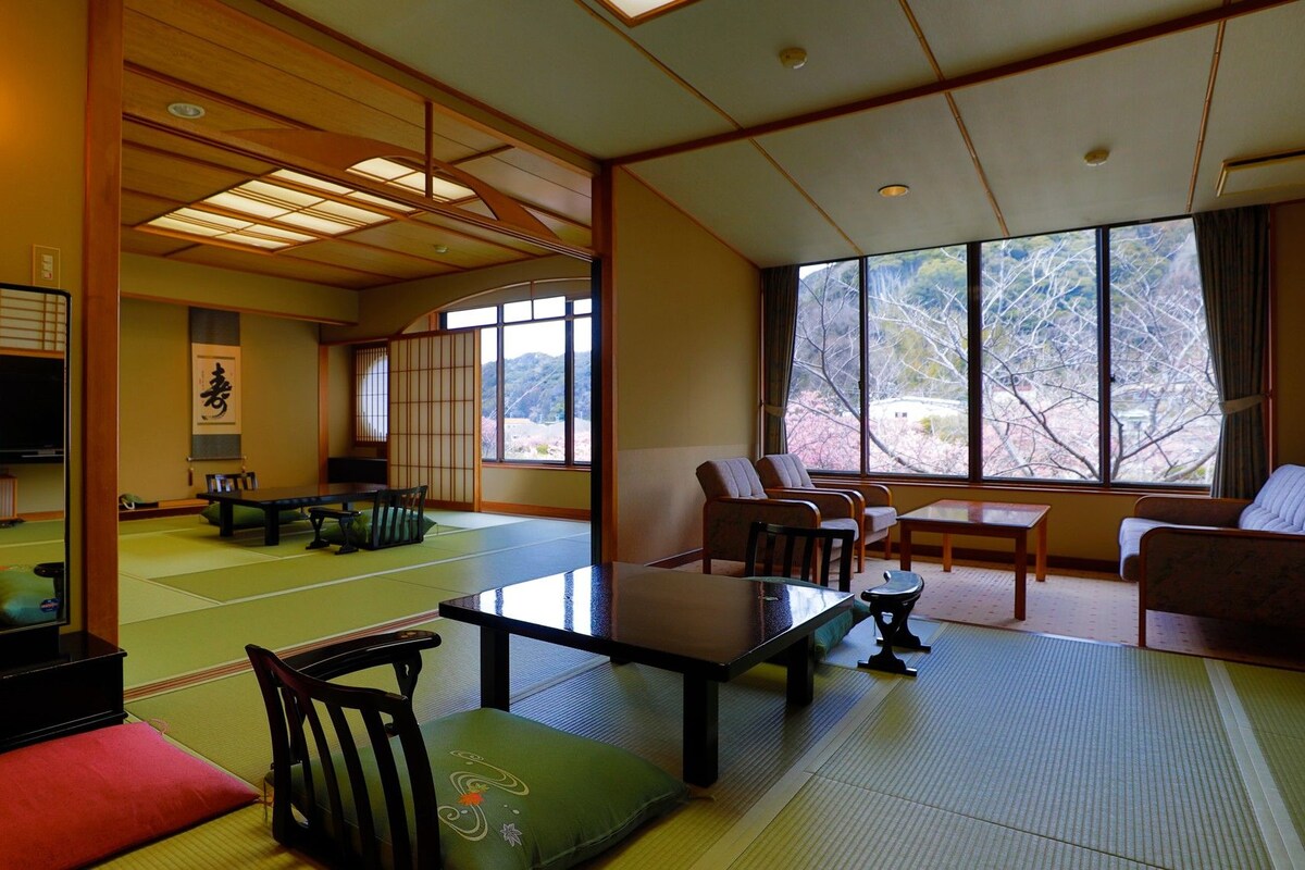 Una habitación japonesa tradicional con suelo de tatami, una mesa baja, sillas y un ventanal con vistas a árboles.