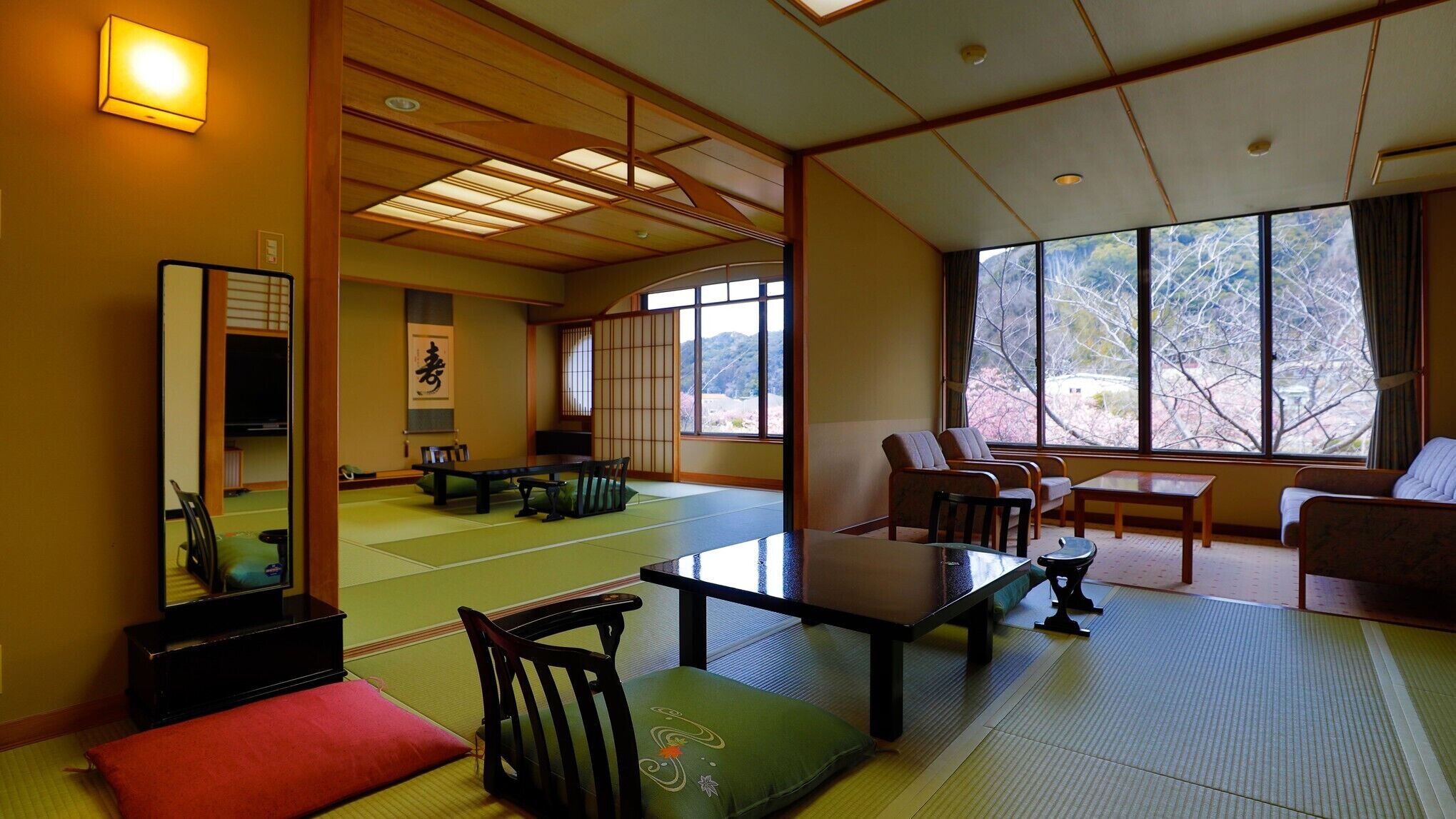 Ein traditioneller japanischer Raum mit Tatami-Böden, einem niedrigen Tisch, Stühlen und einem großen Fenster mit Blick auf Bäume.