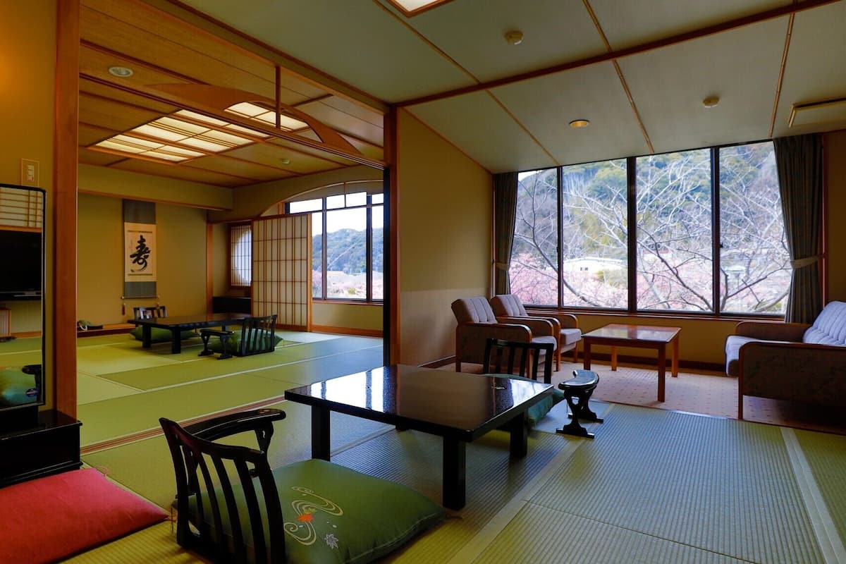 A traditional Japanese room with tatami flooring, a low table, chairs, and a large window offering a view of trees.