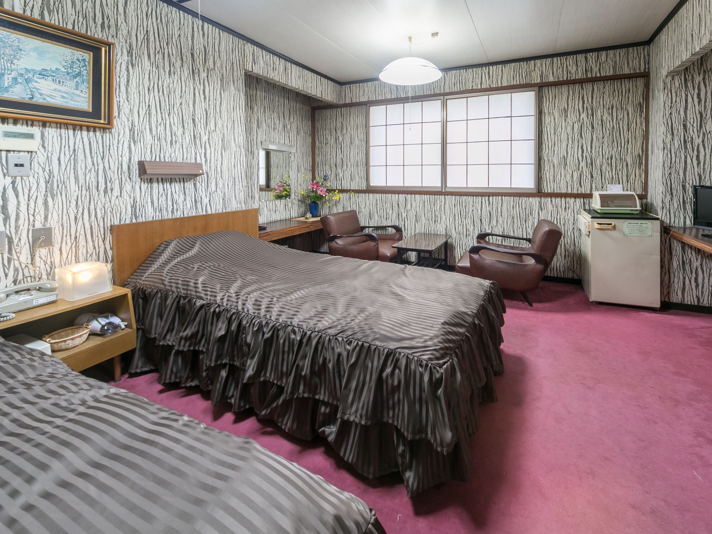 A hotel room with two beds, a desk, a chair, a small refrigerator, and a window with a view.