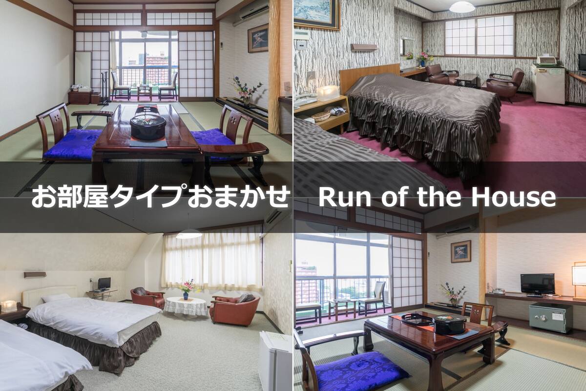 A traditional Japanese-style hotel room with tatami flooring, a tatami mat table, a bed with a futon, a small balcony, and a view of the city.