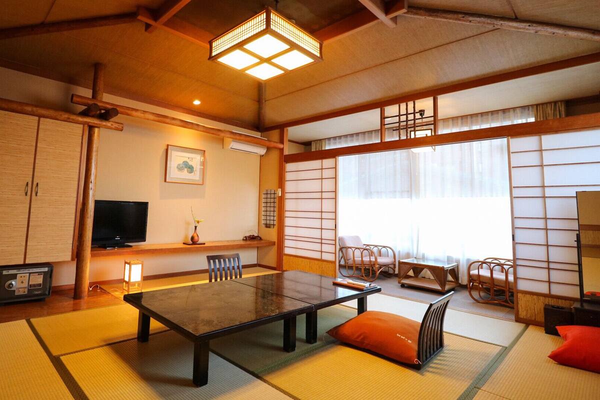 Una habitación tradicional japonesa con suelo de tatami, una mesa baja, sillas, un televisor y puertas correderas.