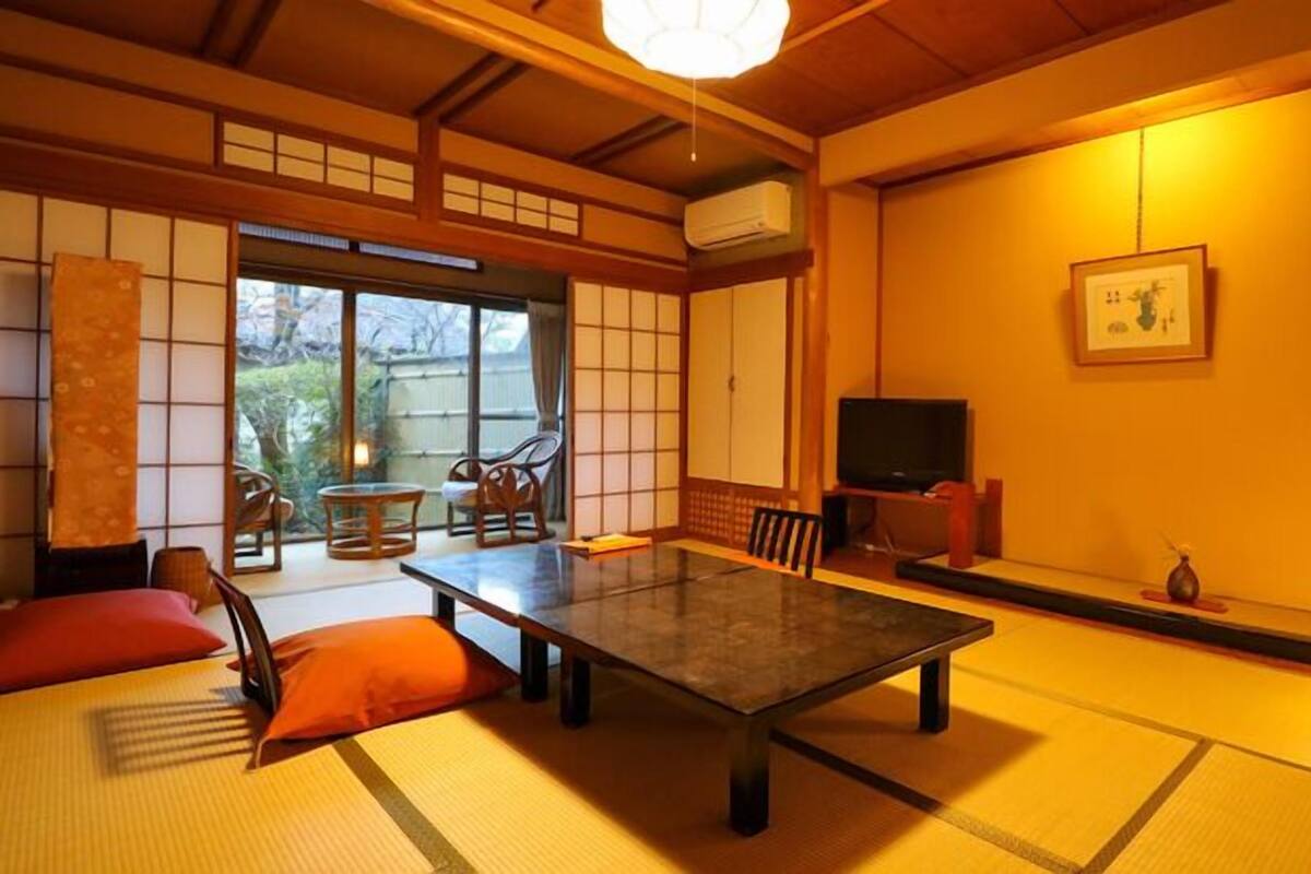 Una habitación japonesa tradicional con suelo de tatami, una mesa baja, sillas, un televisor y una puerta corredera que da a un jardín.