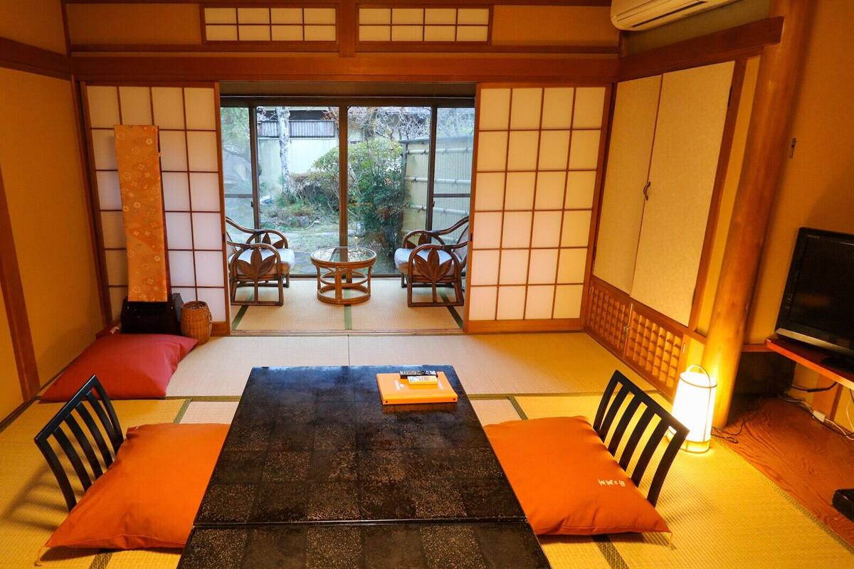 Una habitación japonesa tradicional con suelo de tatami, una mesa baja, cojines, un televisor y vistas a través de un puertas correderas.