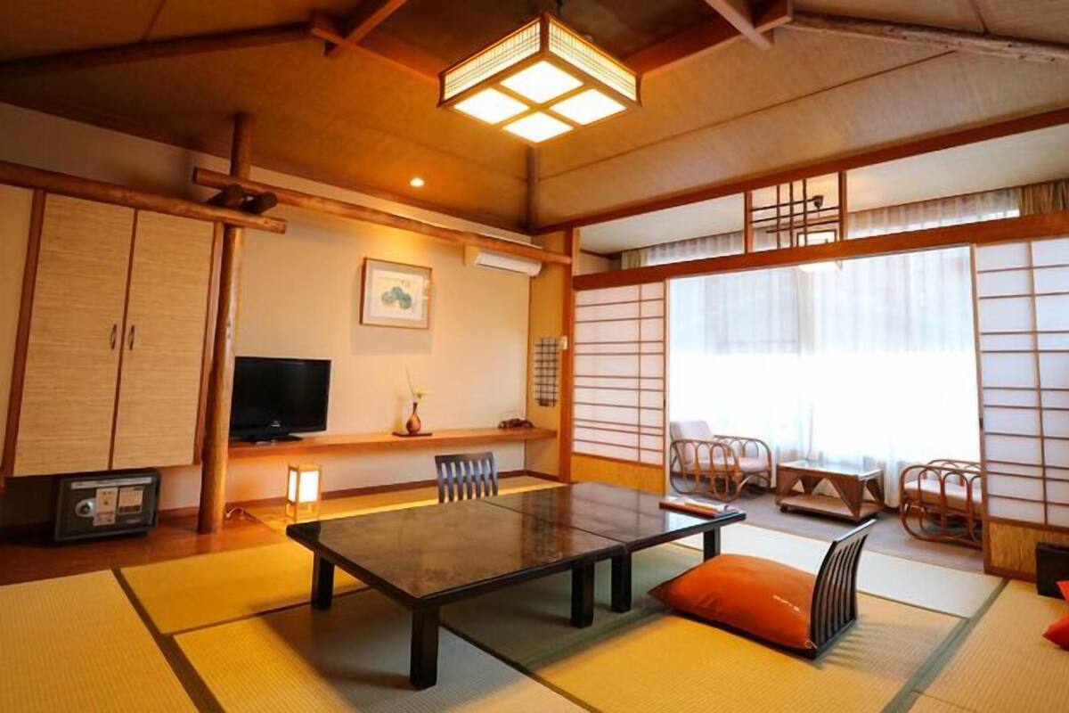 Una habitación japonesa tradicional con suelo de tatami, una mesa baja, sillas, un televisor y puertas correderas.
