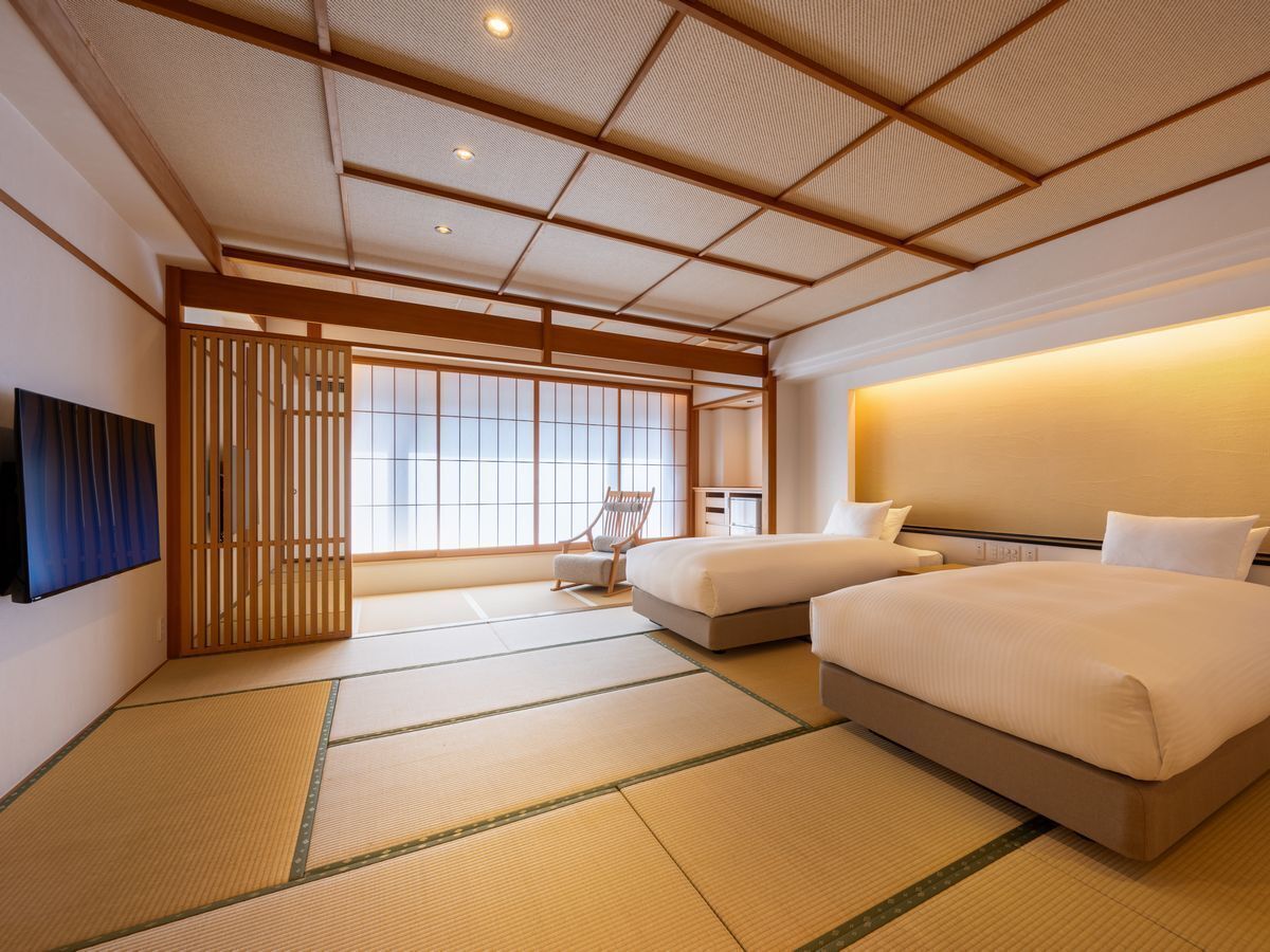 A traditional Japanese-style hotel room with tatami flooring, a low ceiling with wooden beams, a sliding door, and a bed with white bedding.