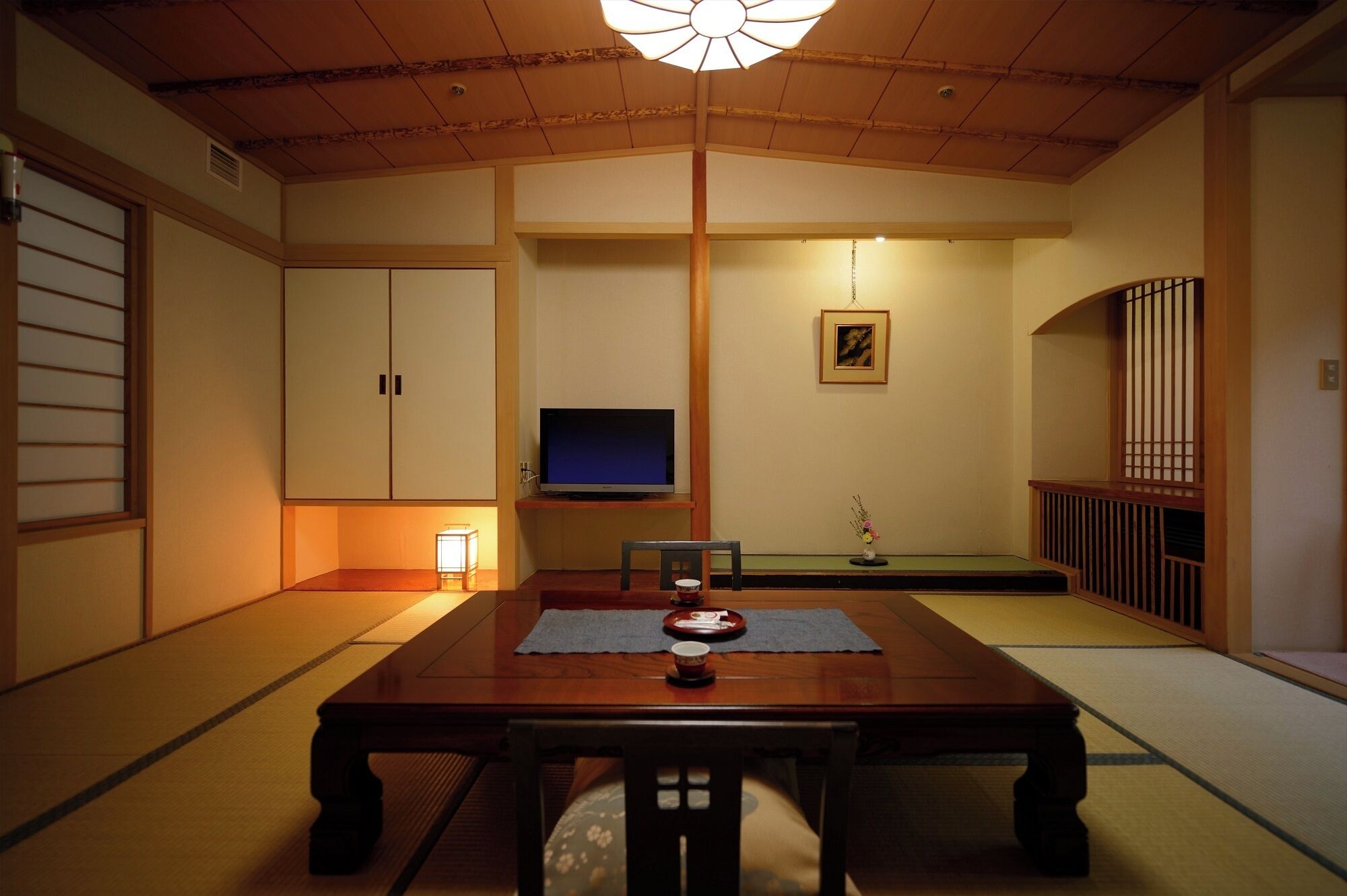 A traditional Japanese room with tatami flooring, a low table with a cushion, a television on a stand, and a sliding door.