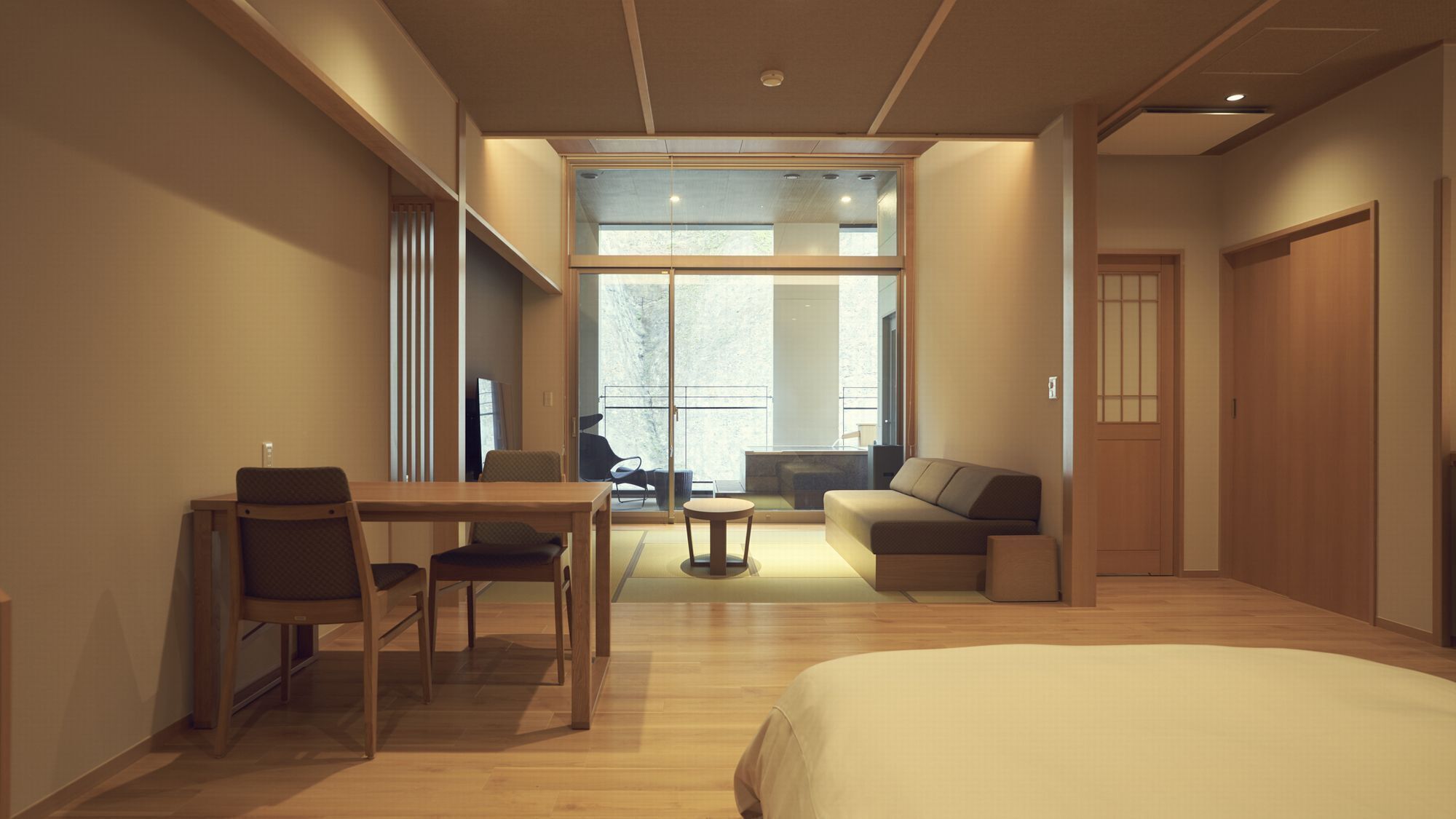 A hotel room with a bed, a desk with chairs, a sofa, and a balcony with a view.
