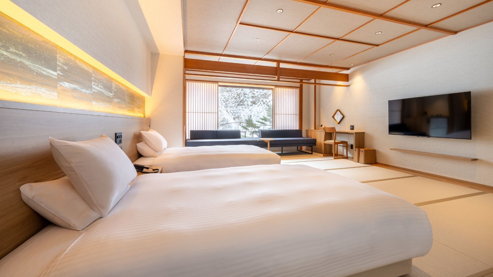 A modern hotel room with two beds, a TV, a desk, and a view of trees through the window.