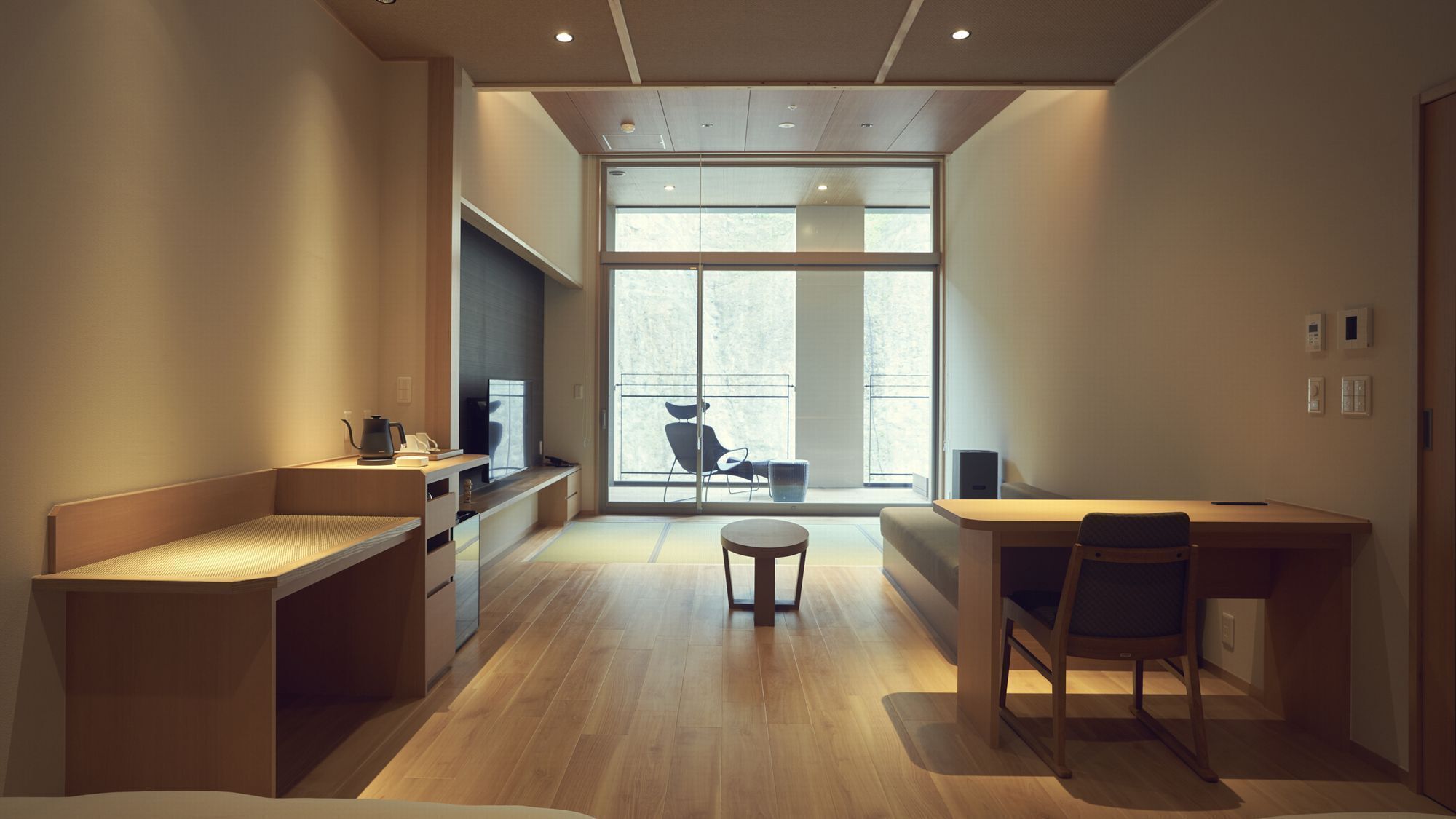 A modern, minimalist room with a wooden desk, a chair, a small table, and a sofa. There is a large window providing natural light and a view of the outdoors.