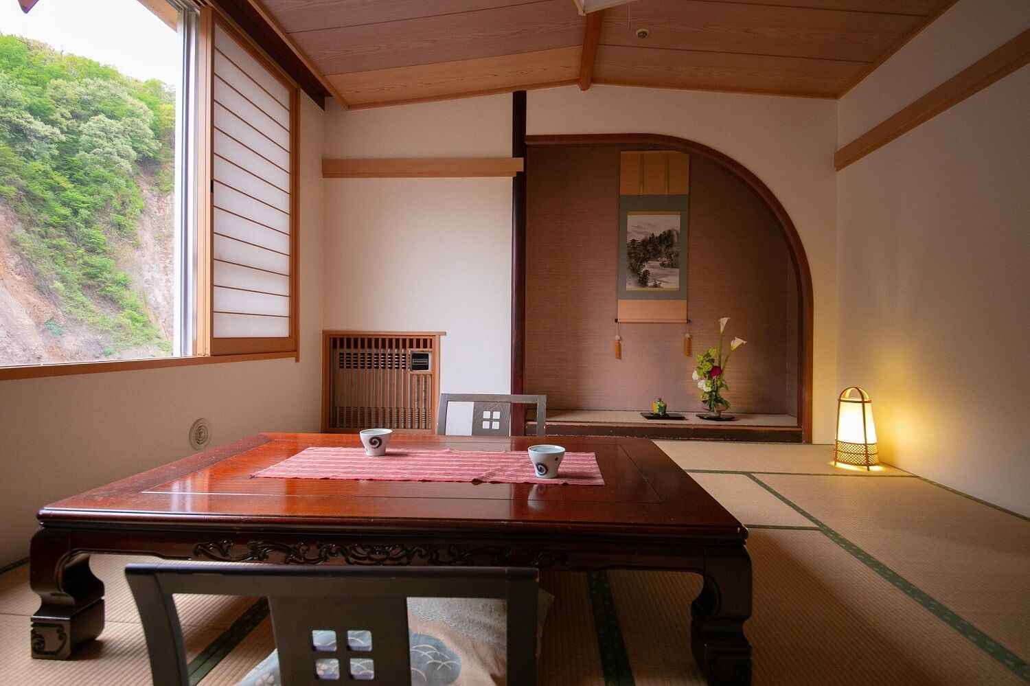 A traditional Japanese room with tatami flooring, a wooden table, and a sliding door leading to a view of a mountain.