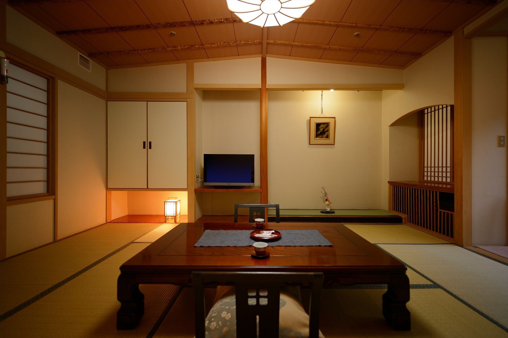 A traditional Japanese room with tatami flooring, a low table with a cushion, a television, and a painting on the wall.