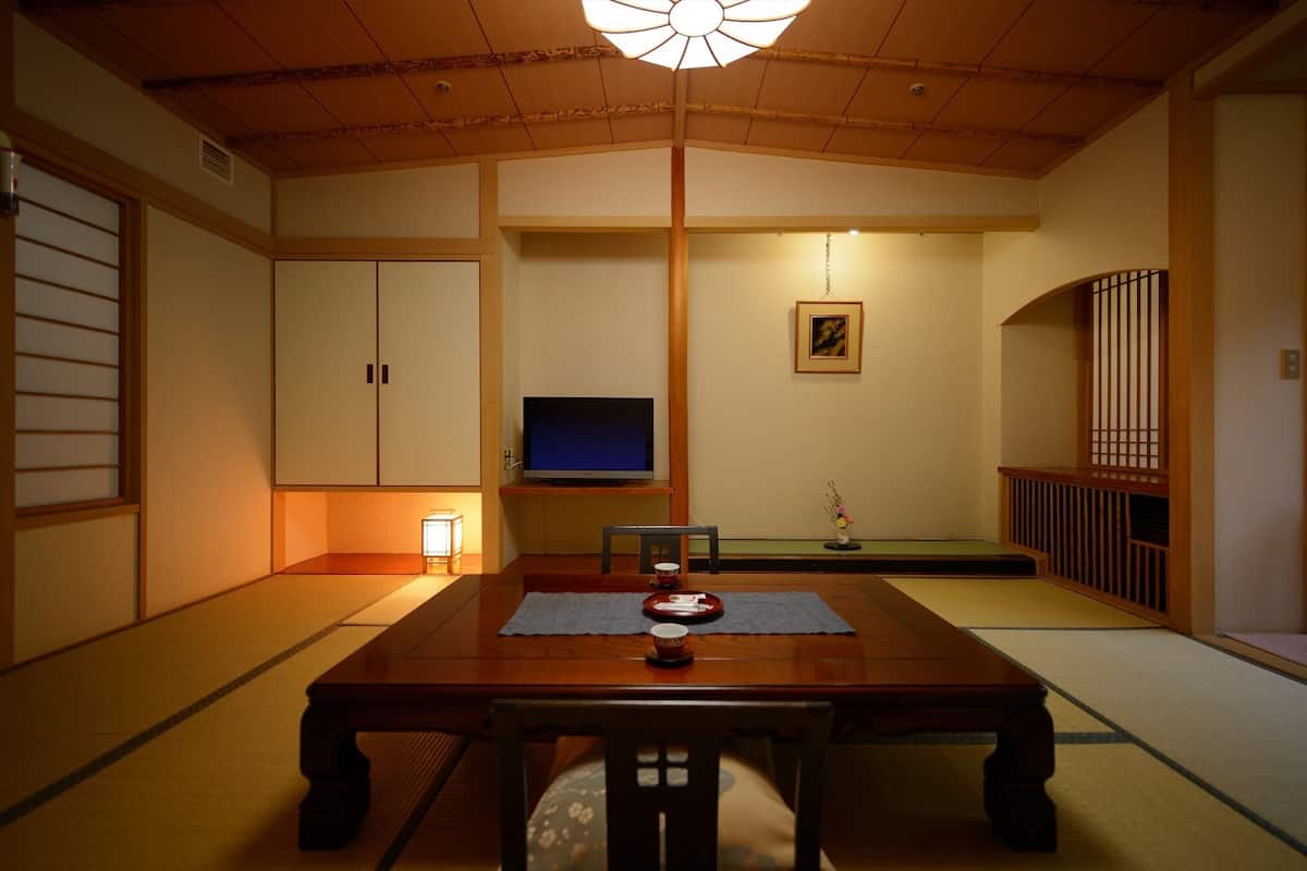 A traditional Japanese room with tatami flooring, a low table with a cushion, a television, and a painting on the wall.