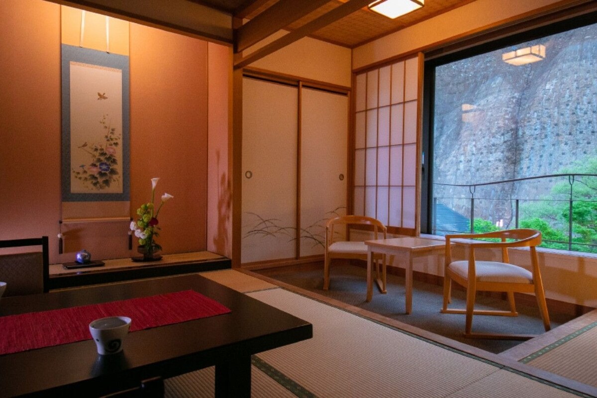 A traditional Japanese room with tatami flooring, a low table with a red placemat, and a view of a mountainous landscape through a sliding door.