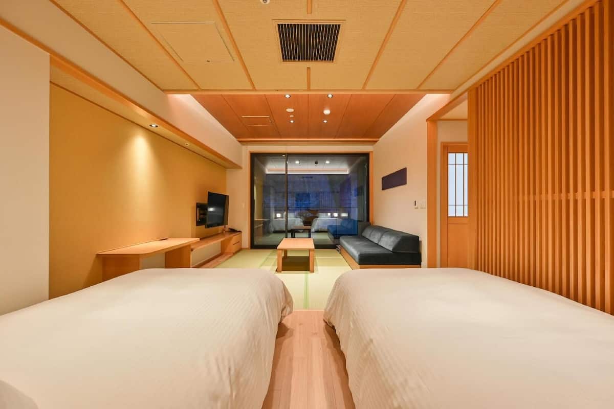 A hotel room with two beds, a TV, a sofa, and a sliding glass door leading to a balcony.