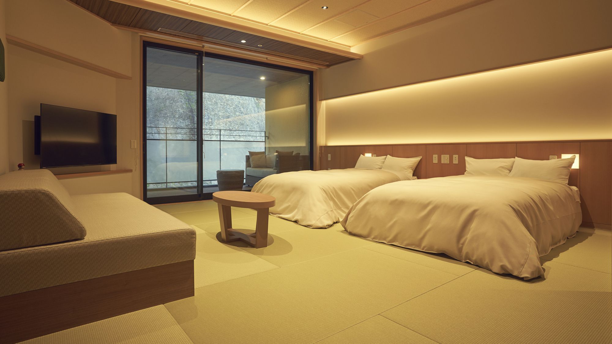 A hotel room with two beds, a sofa, a small table, a TV, and a sliding glass door to the outside.