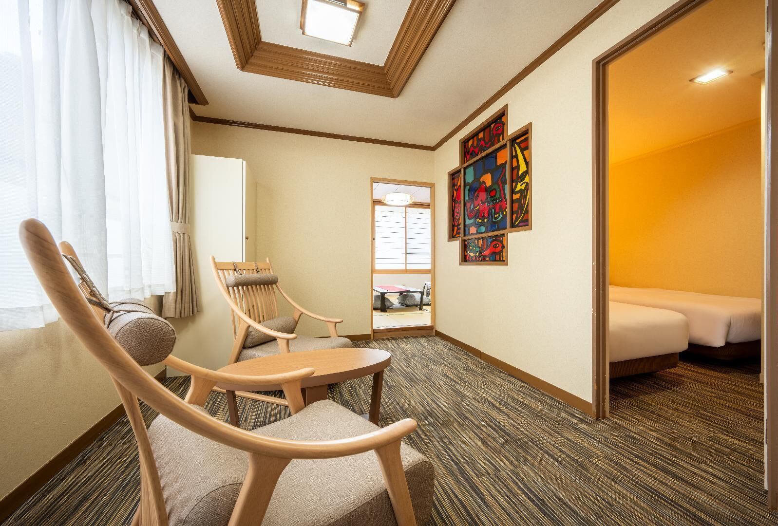 A hotel room with a bed, a chair, a table, a window with curtains, and a wall with colorful artwork.