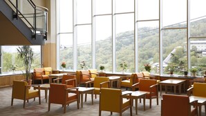 A spacious indoor area with multiple tables and chairs, large windows offering a view of a forested hillside, and a staircase leading to an upper level. - Manza Hotel Juraku (Tsumagoi)