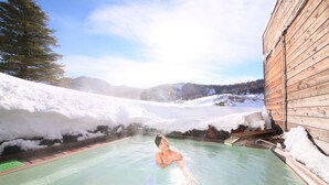 A person in a hot spring surrounded by snow and a wooden building. - Manza Hotel Juraku (Tsumagoi)