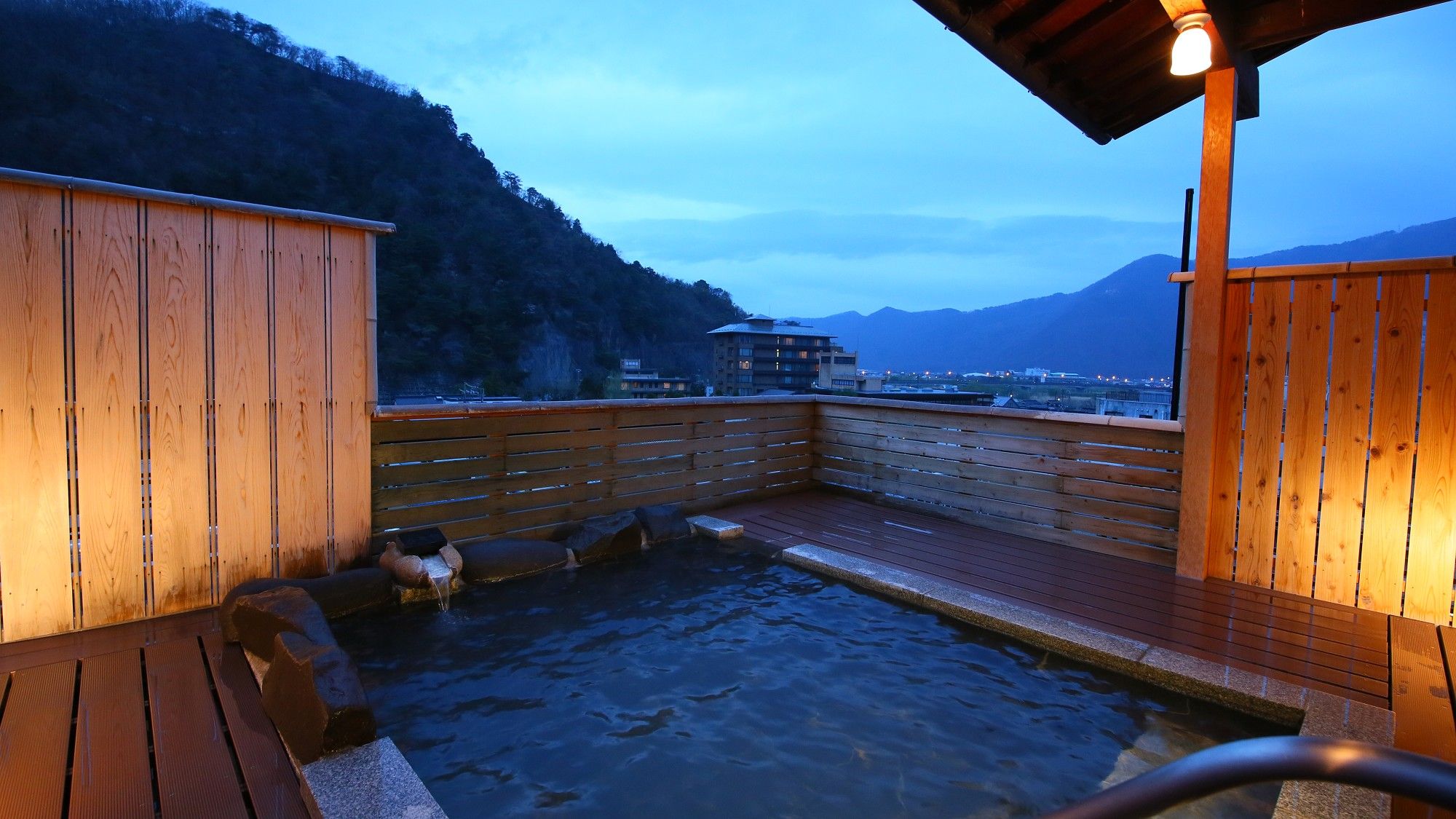 Una terraza de madera con un baño de aguas termales, bancos de madera y vistas a montañas y edificios al fondo.