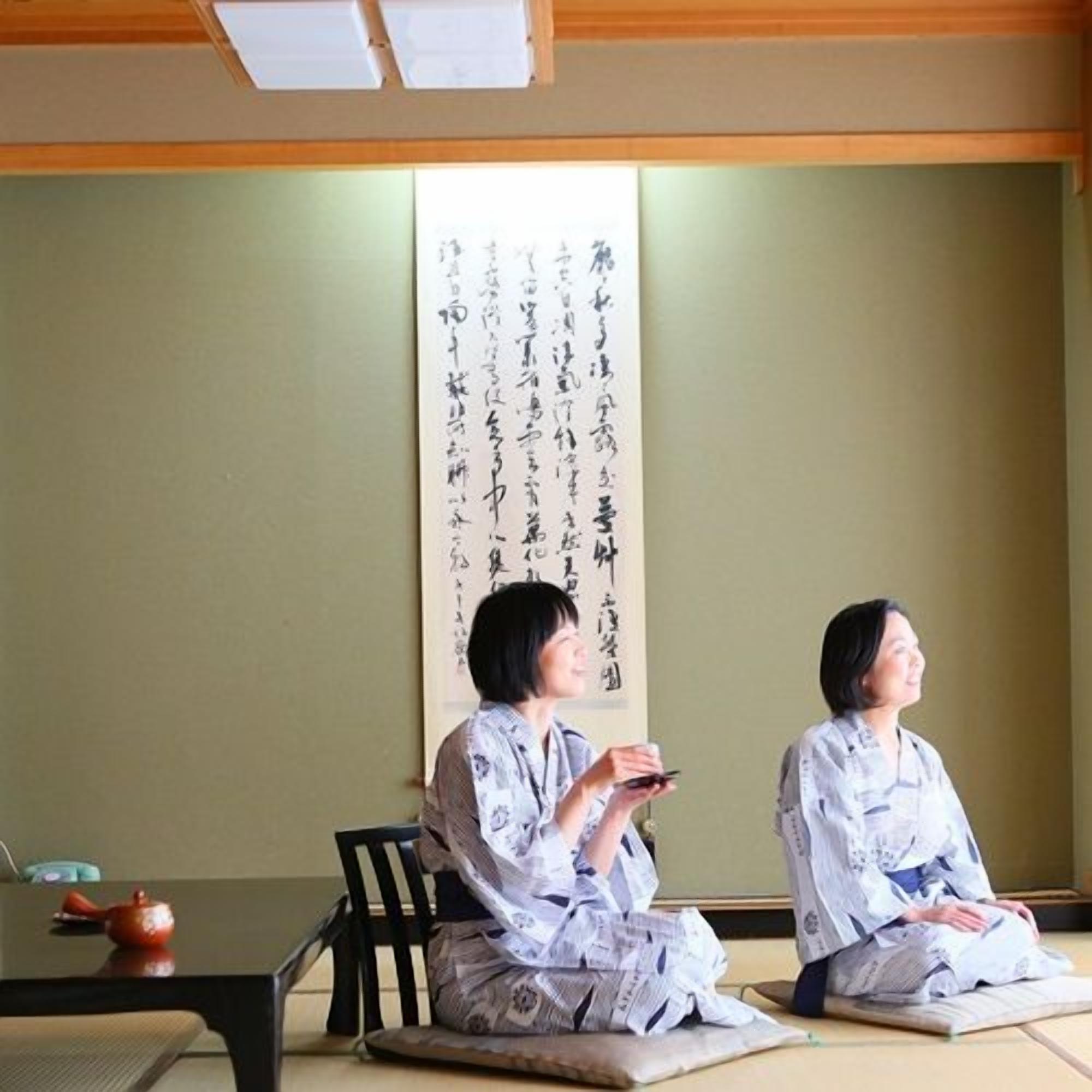 Dos personas sentadas sobre esteras de tatami en una habitación japonesa tradicional con un biombos shoji y un rollo de caligrafía en la pared.