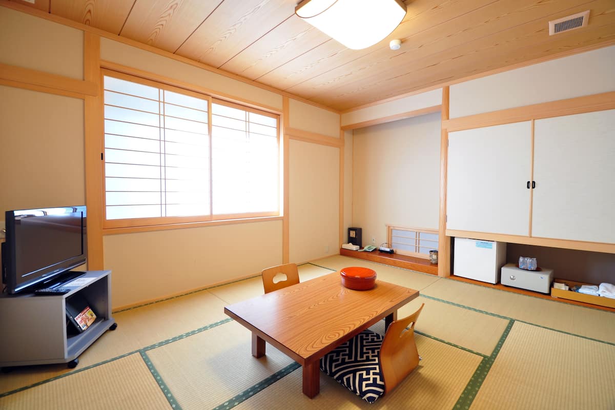 A traditional Japanese room with tatami flooring, a low table with a red bowl, a television on a shelf, and sliding doors.