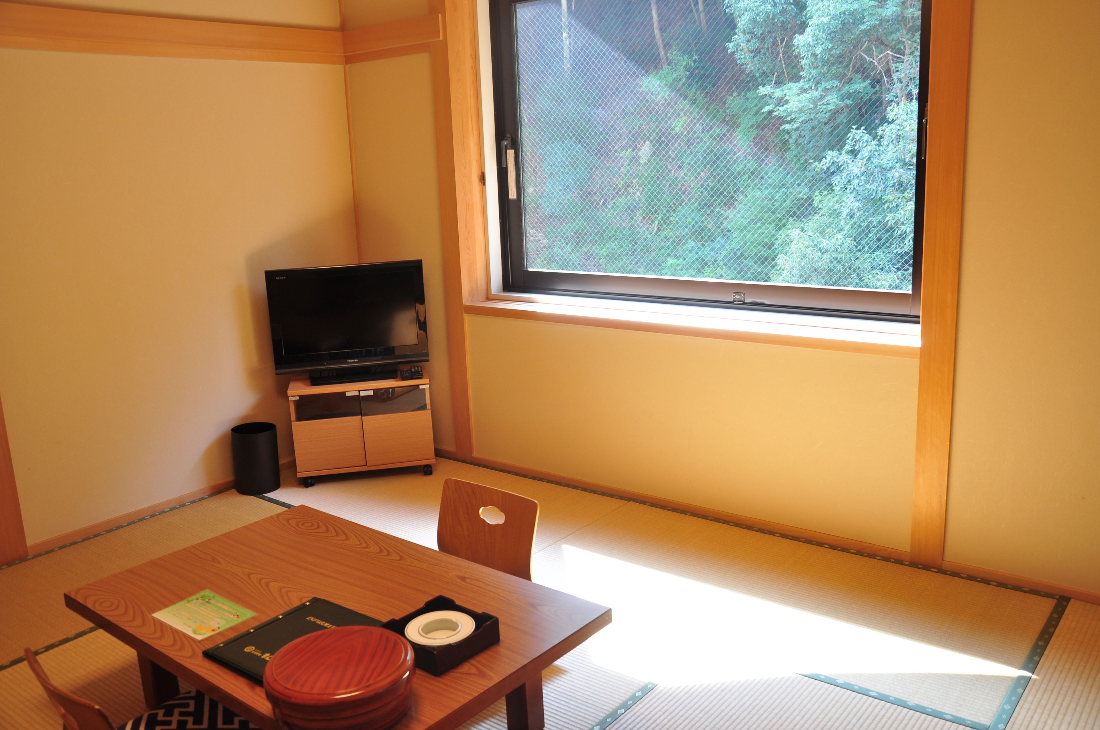 A traditional Japanese room with tatami flooring, a low table with a red stool, a television on a wooden stand, and a large window with a view of greenery.