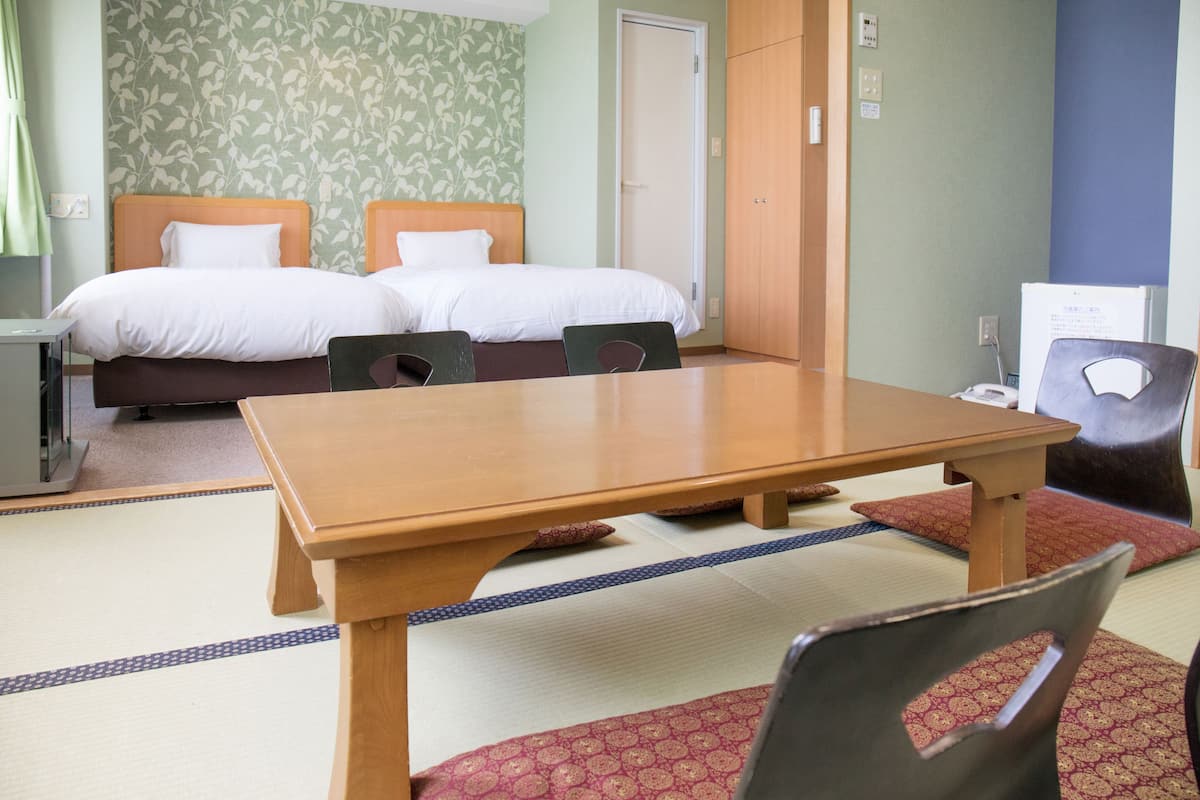 A hotel room with two beds, a wooden table, chairs, a desk, and a small refrigerator.