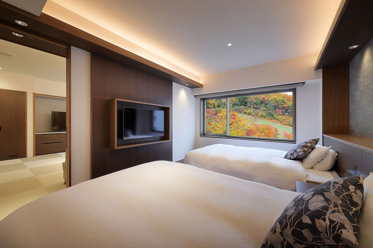A hotel room with two beds, a TV, a window with a view of autumn foliage, and a sliding door closet.