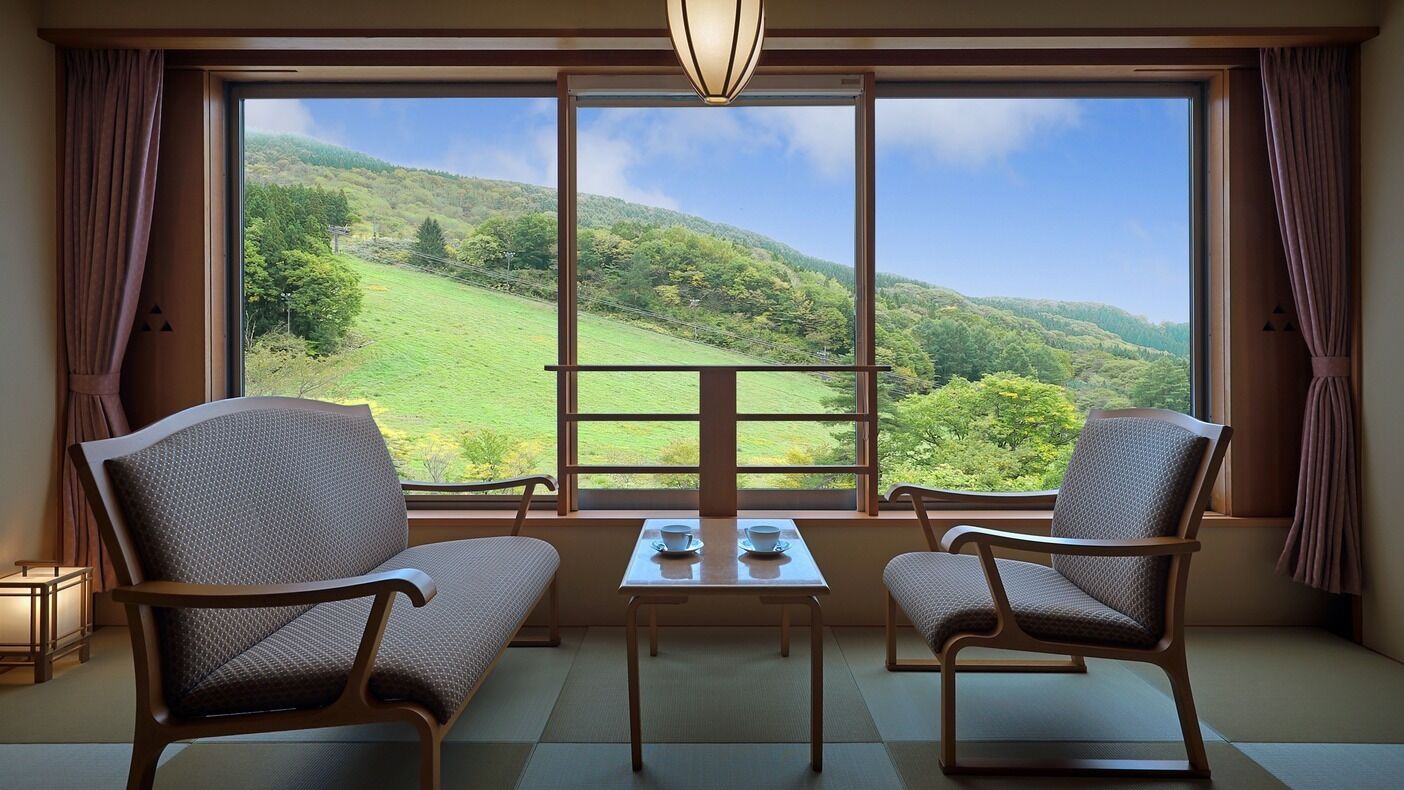A room with two chairs, a table, and a view of a green landscape through large windows.