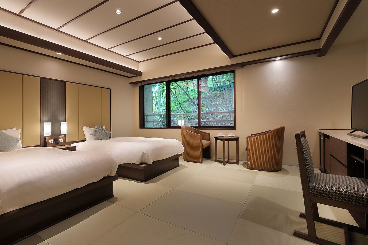 A modern hotel room with two beds, a desk, a chair, and a large window offering a view of trees.