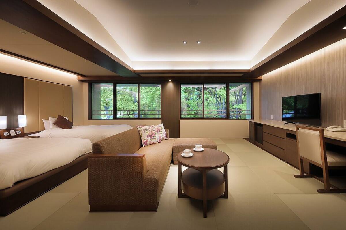 Una habitación de hotel moderna con una cama grande, un sofá, una mesa redonda, un escritorio, un televisor y ventanas amplias con vistas a árboles.