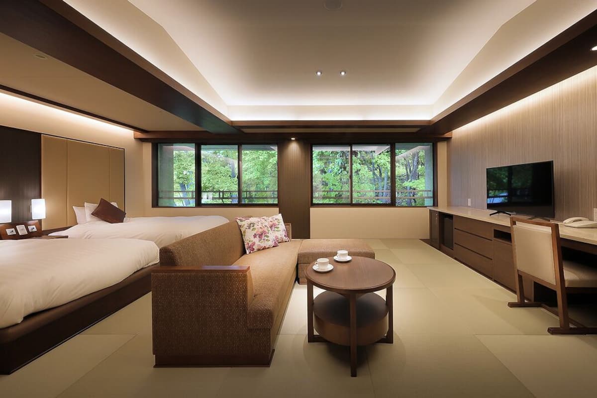A modern hotel room with a large bed, a sofa, a round table, a desk, a TV, and large windows with a view of trees.