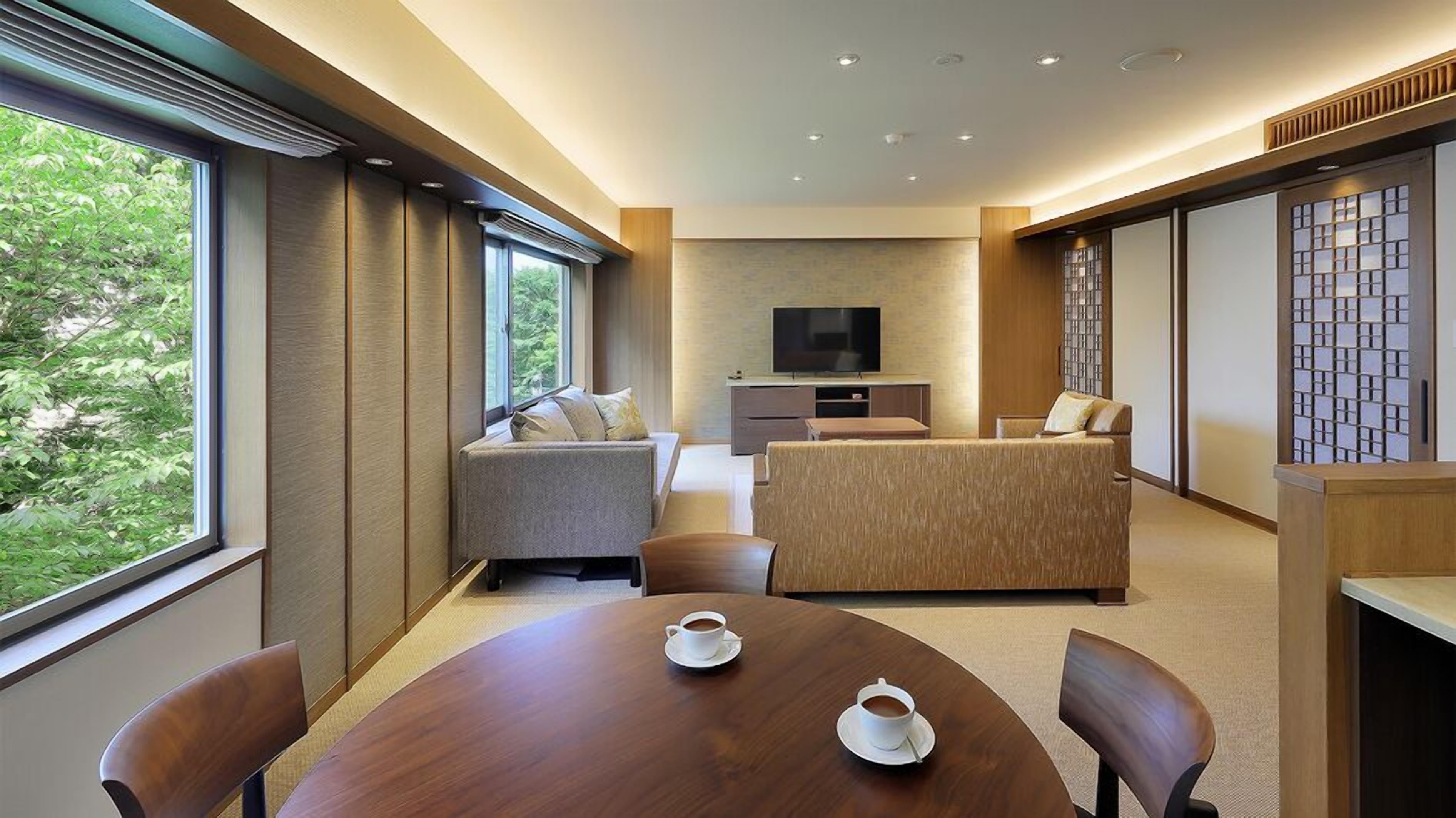 A modern hotel room with a sofa, a TV, a dining table with two cups, and a view of greenery outside.