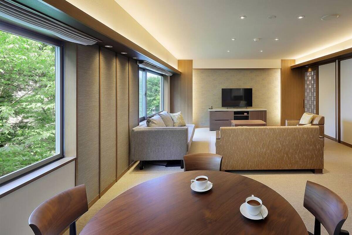 Habitación de hotel moderna con sofá, televisión, mesa de comedor con dos tazas y vistas a zonas verdes.