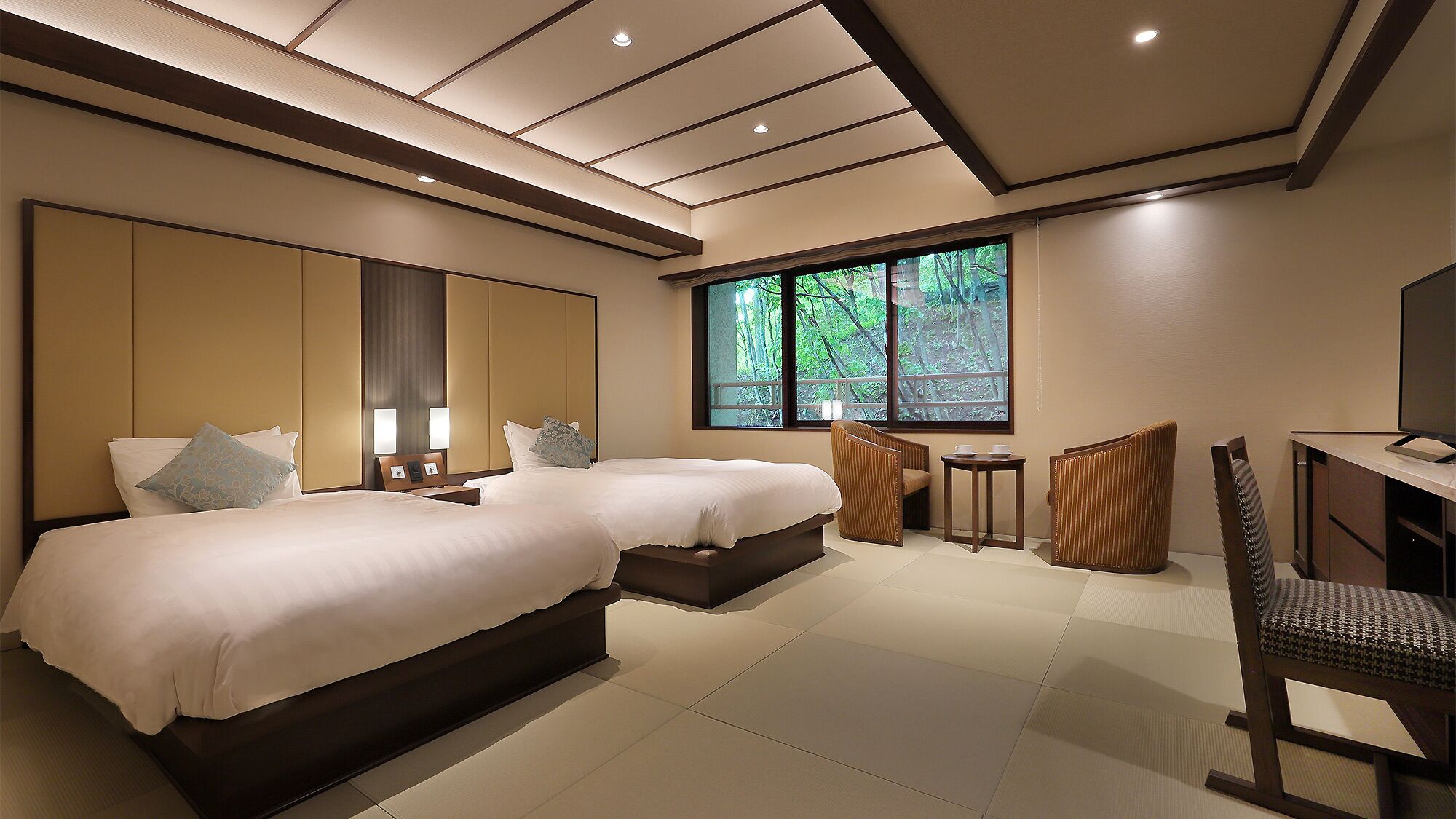 A hotel room with two beds, a desk, a chair, and a large window with a view of trees.