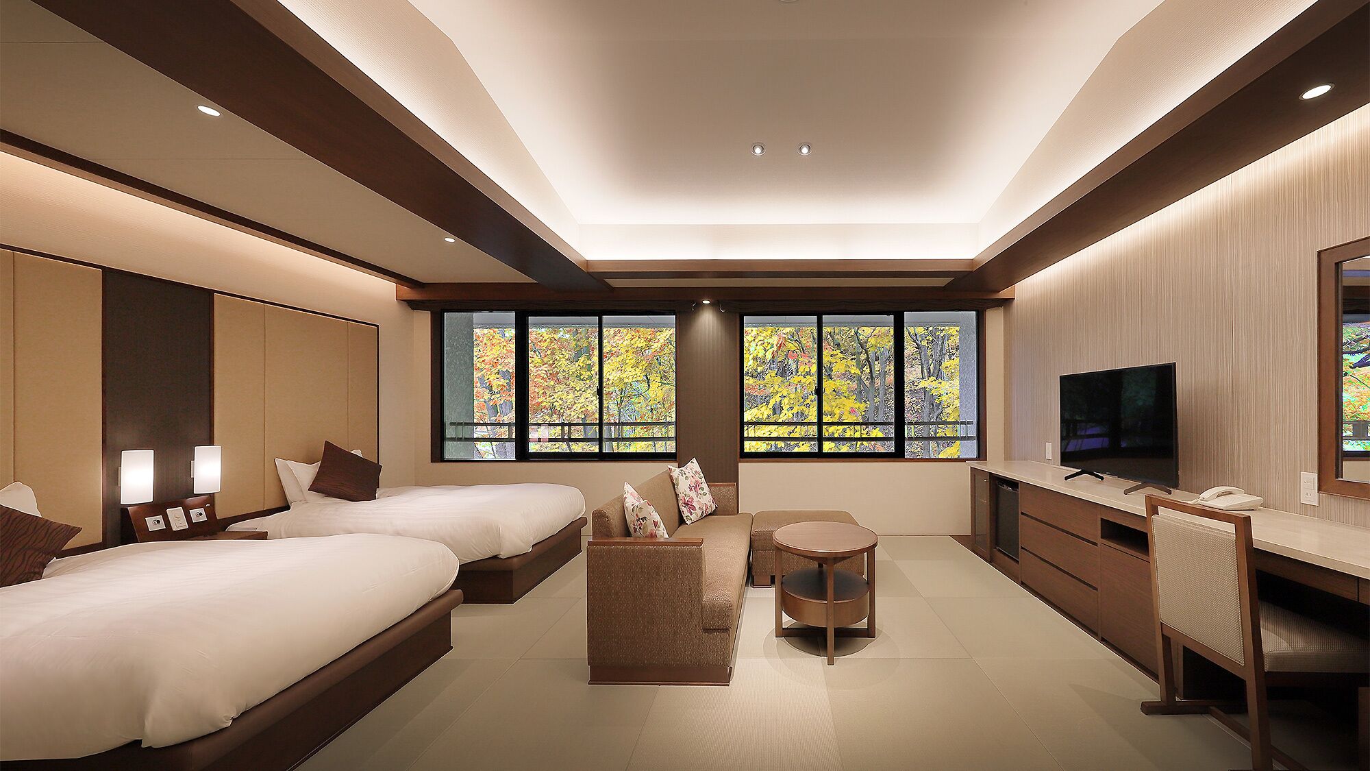Habitación de hotel con dos camas, un escritorio, un televisor y ventanas amplias con vistas al follaje otoñal.