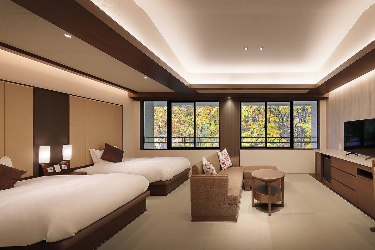 Habitación de hotel con dos camas, un escritorio, un televisor y ventanas amplias con vistas al follaje otoñal.