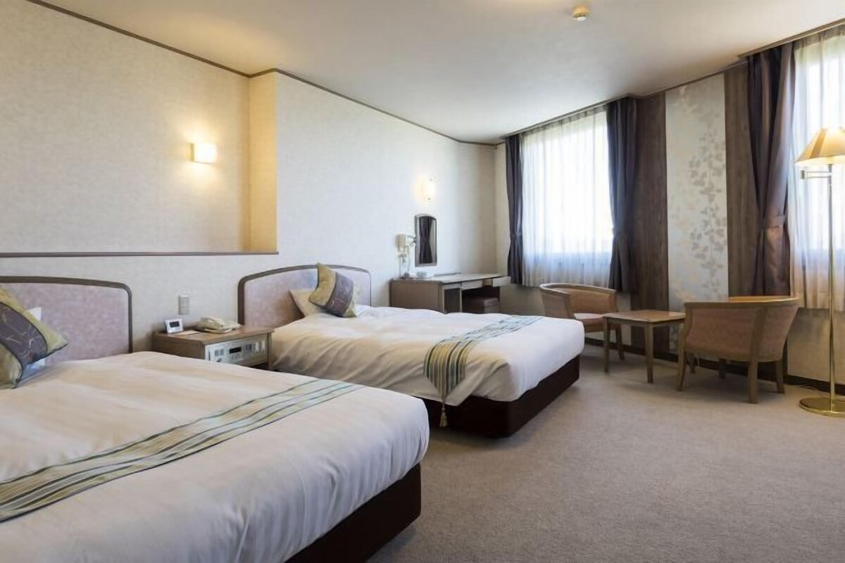 A hotel room with two beds, a desk, a chair, a lamp, and a window with curtains.