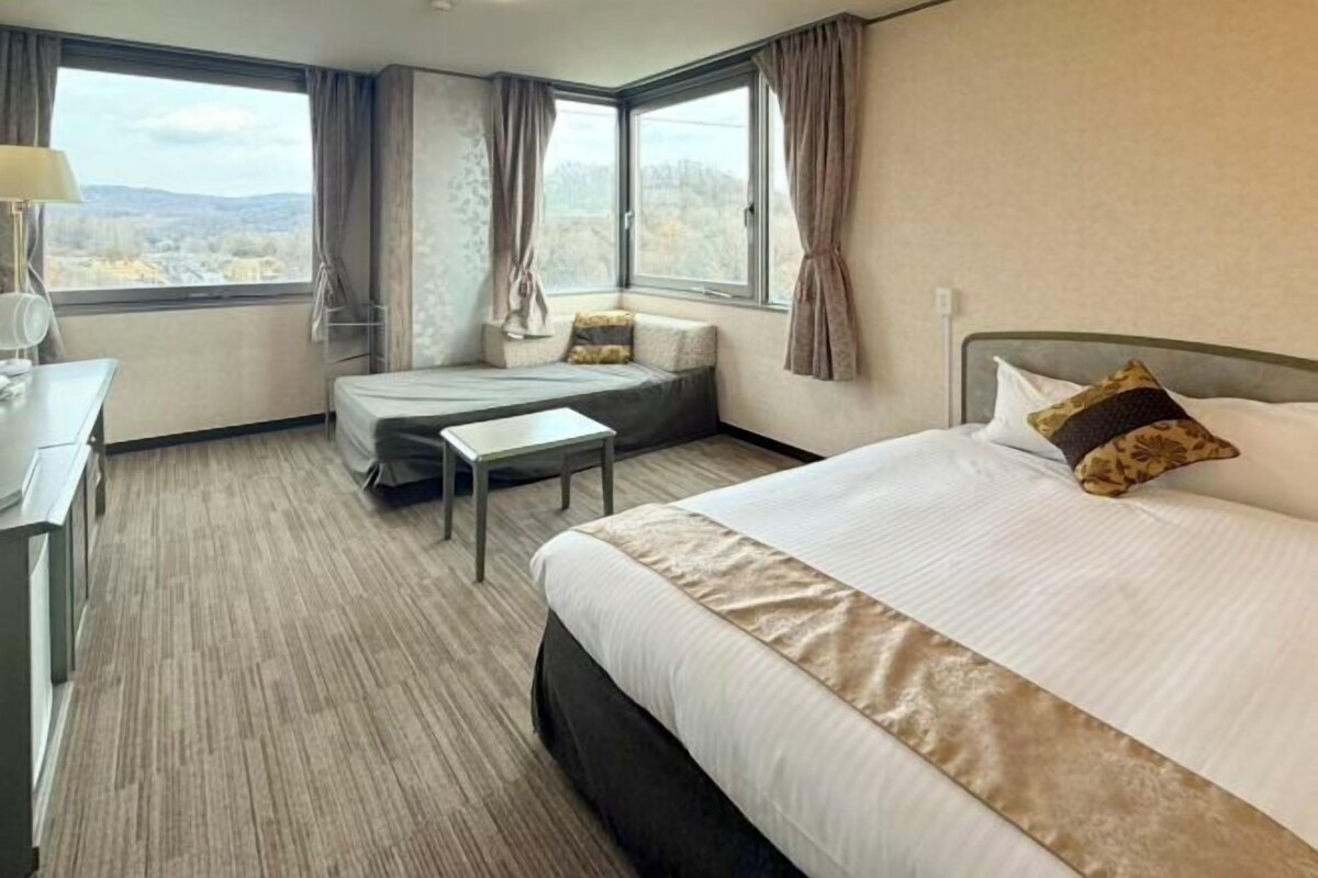 A hotel room with a bed, a desk, a chair, and a view of the outdoors through large windows.