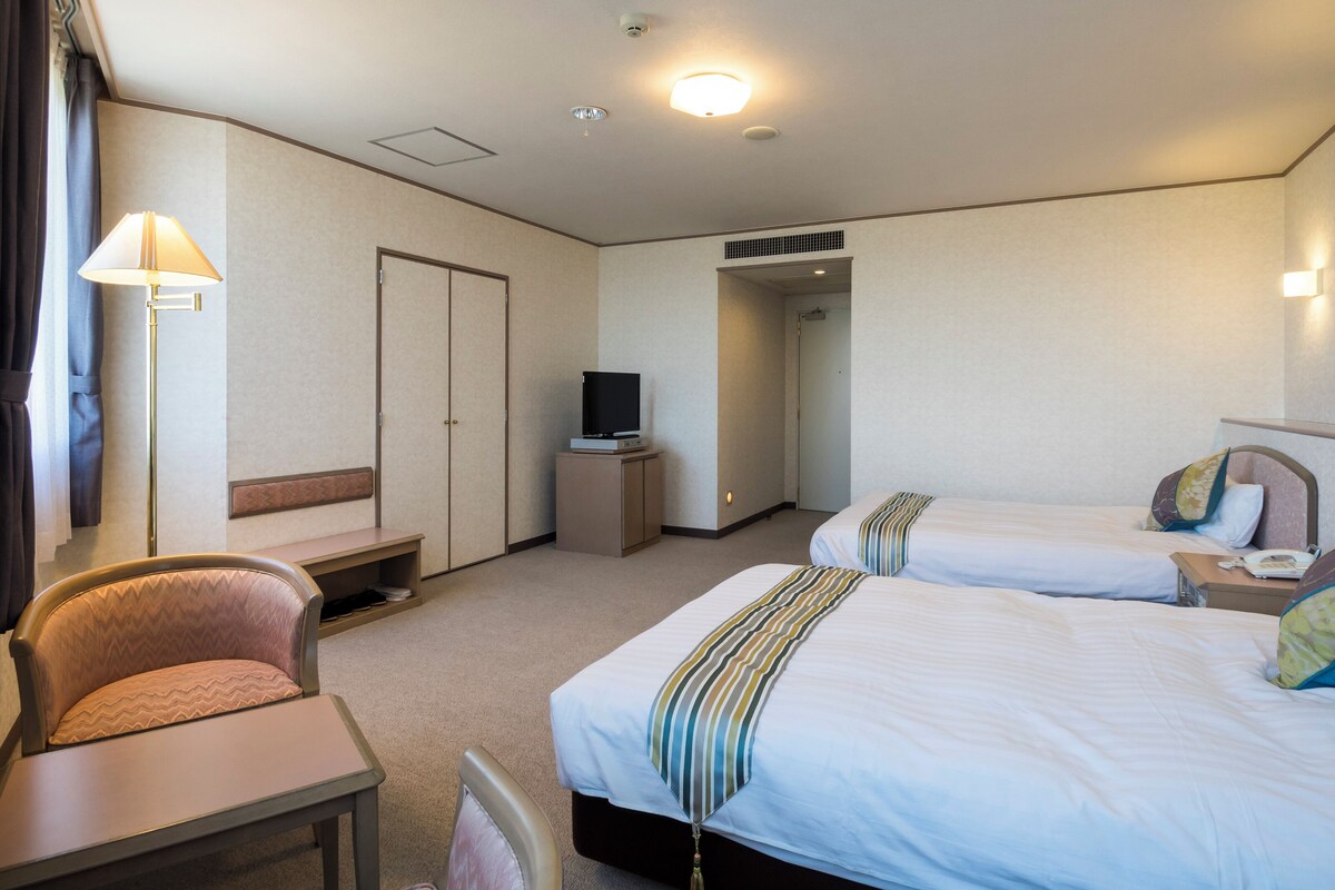 A hotel room with two beds, a desk, a chair, a television, and a lamp.