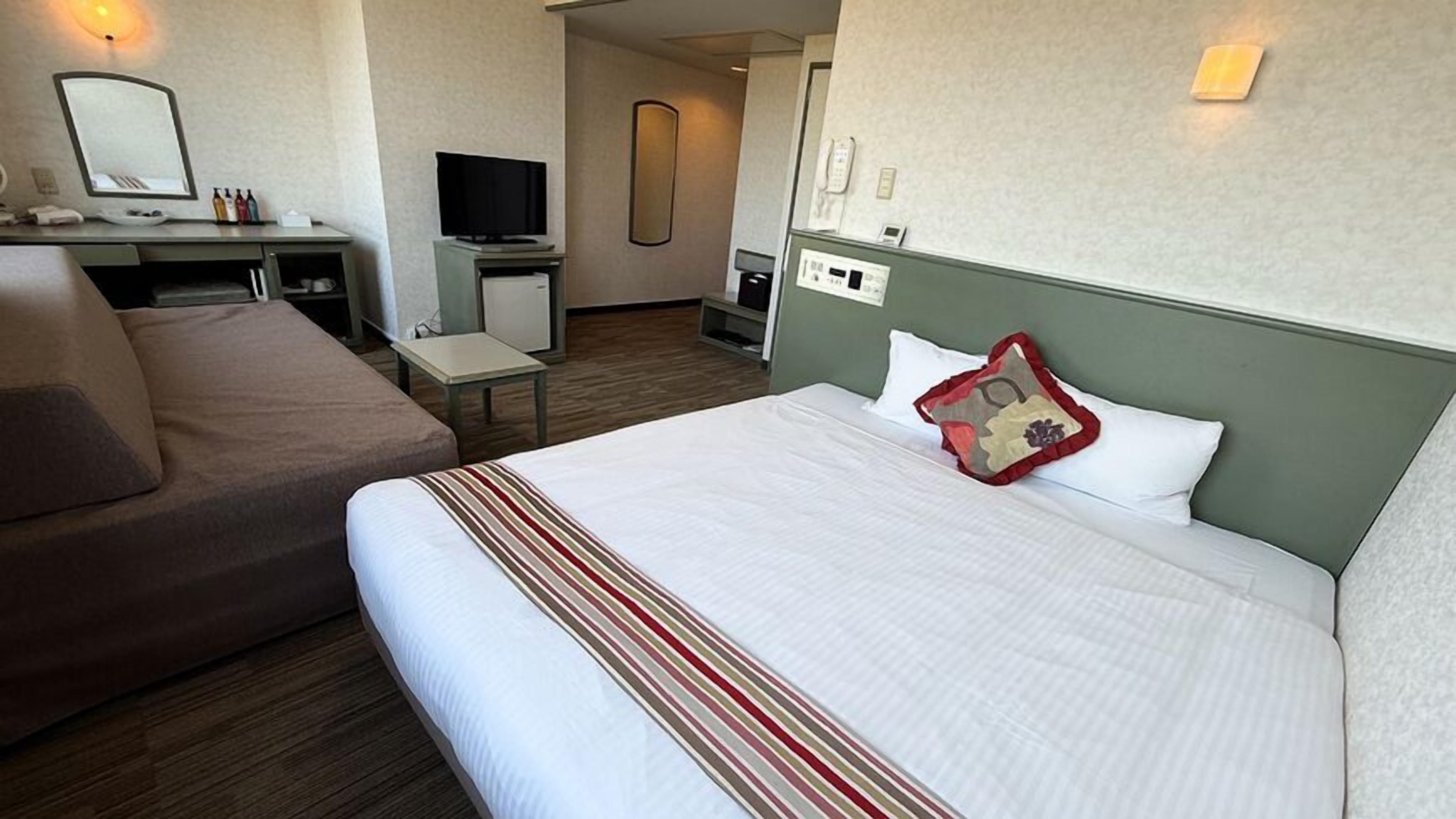 A hotel room with a bed, a sofa, a desk with a TV, a mirror, and a phone.