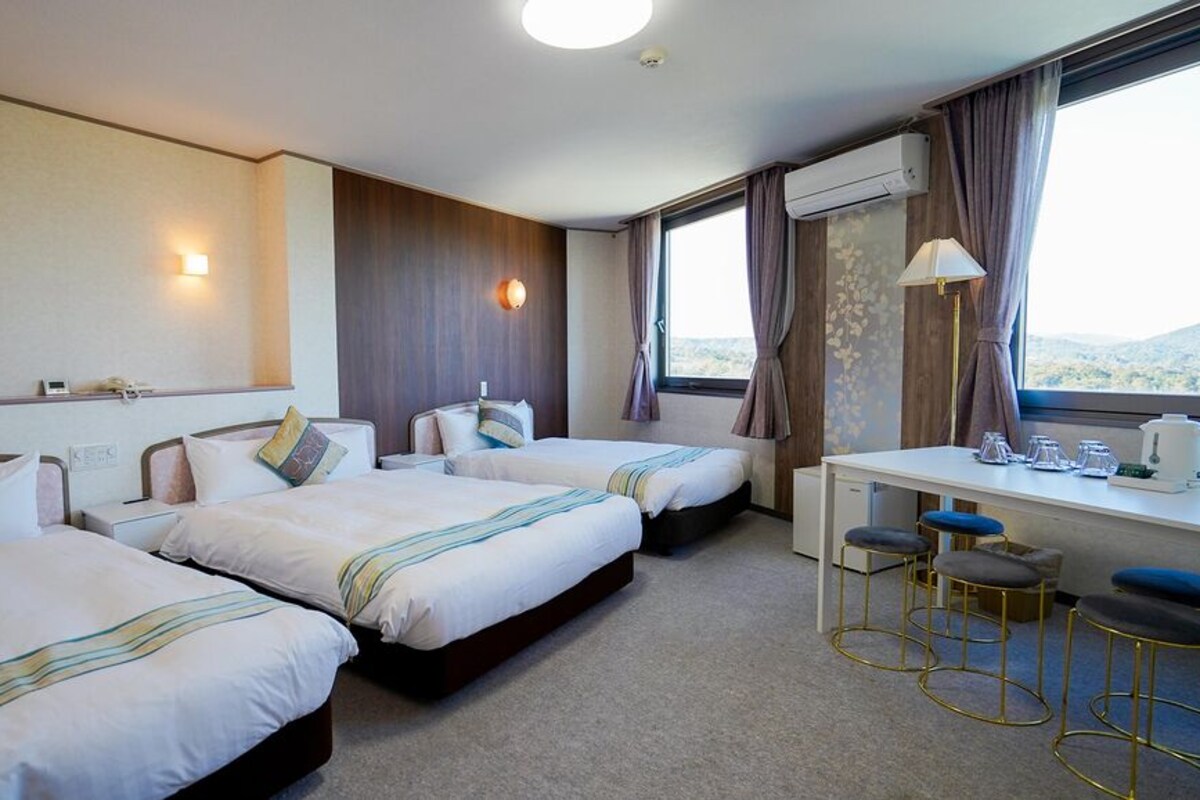 A hotel room with three beds, a desk with bar stools, a window with curtains, and a view of the outdoors.