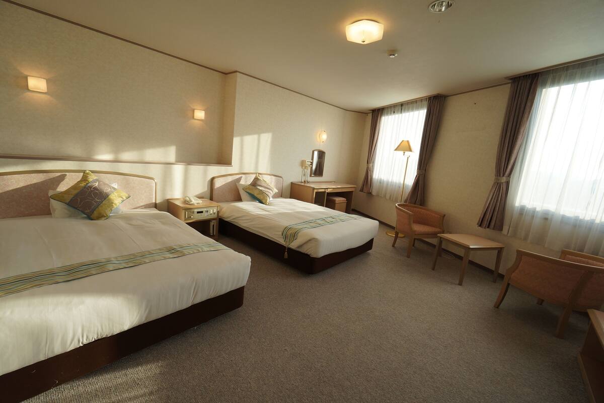 A hotel room with two beds, a desk, a chair, and a lamp.