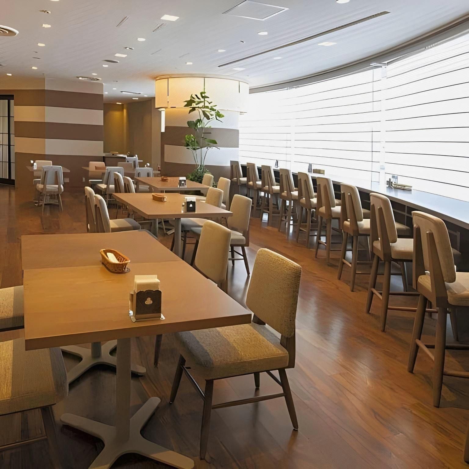 A spacious dining area with multiple tables and chairs, a large window with blinds, and a plant in the center.